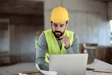 Middle-aged male with dark beard and mustache and wearing a hard had and yellow safety vest while sitting at a table in front of a laptop in a construction zone. 